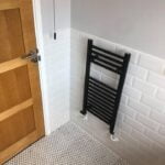 Modern bathroom installation showing a white tiled wall, a black towel radiator, and a wooden door.