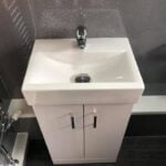 A modern white bathroom sink with a chrome faucet, set against a gray speckled wall with bathroom tiling and a cabinet underneath.