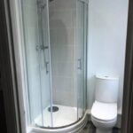 A modern disabled bathroom corner shower cubicle next to a white toilet in a bathroom with gray tiles.
