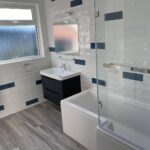 A modern disabled bathroom interior with a white and blue tile design, including a sink, a mirror, and a bathtub with a glass partition.