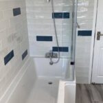 Modern bathroom with a white bathtub and grey tiled walls featuring blue accents, designed by a professional bathroom fitter, equipped with a glass shower door.