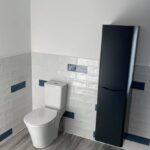 A modern bathroom with white walls, tiled with a blue accent strip, featuring a toilet and a tall, black cabinet, designed for disabled bathroom installation.