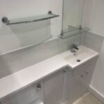 A modern, disabled bathroom with a white vanity cabinet, rectangular basin, chrome faucet, and a glass shelf atop a tiled backsplash.