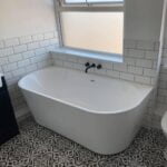 A modern freestanding bathtub in a bathroom with black-and-white patterned floor tiles, white subway wall tiles, and accommodations for the disabled.