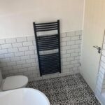 A black towel radiator, expertly installed by a bathroom fitter, mounted on a white tiled wall in a bathroom with patterned floor tiles.