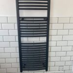 A black towel radiator mounted on a white tiled wall in a disabled bathroom.