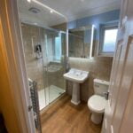 A modern disabled bathroom interior with a glass shower cubicle, pedestal sink, toilet, and heated towel rail.