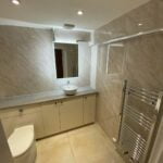 A modern bathroom design with beige tiles featuring a wall-mounted sink, toilet, towel radiator, and a large mirror.