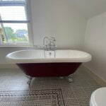 A freestanding white bathtub with a burgundy exterior and silver fixtures in a bright disabled bathroom with patterned tile flooring.