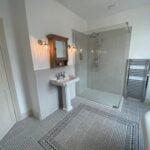 Modern disabled bathroom interior with a glass shower enclosure, pedestal sink, and patterned tile flooring.