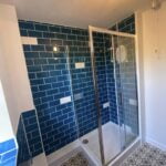 A modern bathroom design with blue subway tiles and a corner shower with glass doors.