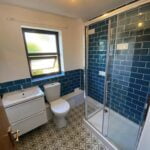 A modern bathroom design with blue tiles, featuring a glass shower enclosure, white sink and toilet, with patterned flooring.