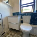 Modern bathroom installation showcasing a white toilet, a vanity with drawers, and blue tiles, complemented by patterned flooring and a frosted window for natural light.