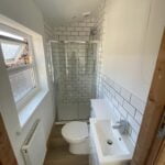 A modern disabled bathroom with white subway tiles, glass shower enclosure, toilet, and a sink with a wood accent wall.