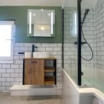 A modern disabled bathroom with white subway tiles, a glass shower partition, and a wooden vanity under a lit mirror.