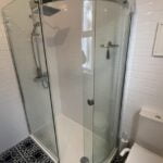 A corner shower with clear glass doors in a disabled bathroom with white subway tiles and patterned floor tiles.