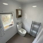 A compact disabled bathroom with a toilet, a wall-mounted cabinet with a mirror, and a heated towel rail, featuring a small window and grey walls.