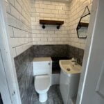 A small, modern disabled bathroom with white subway tiles, dark gray accents, and wooden shelves.