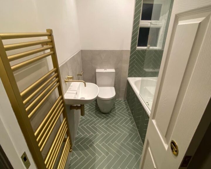 A small disabled bathroom featuring a toilet, a sink, a mirror, and a gold towel radiator, with grey floor tiles and white walls.