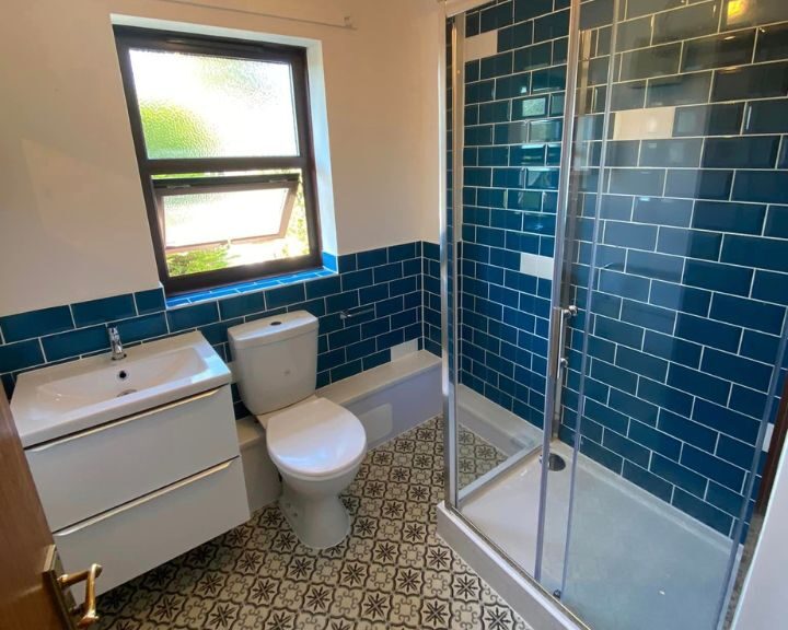 A modern disabled bathroom with blue tiles, featuring a glass shower cubicle, white toilet, sink, and patterned flooring.