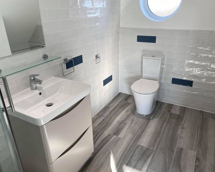Modern bathroom design with white and blue tiles, featuring a sink, toilet, and shower with natural light from a round window.