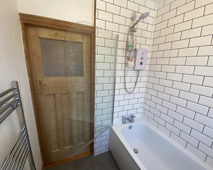 A bathroom corner featuring a white bathtub designed for disabled access, subway tiles on the wall, glass shower partition, electric shower unit, and a wooden door with frosted glass panel.