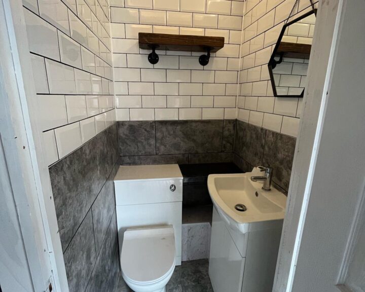 A small, modern disabled bathroom with white subway tiles, dark gray accents, and wooden shelves.