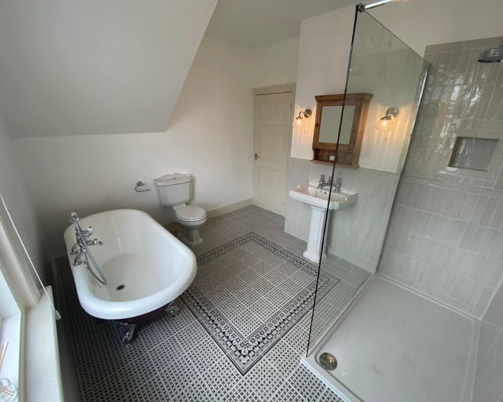 A bright, modern disabled bathroom with a freestanding bathtub, glass shower enclosure, pedestal sink, and patterned floor tiles.